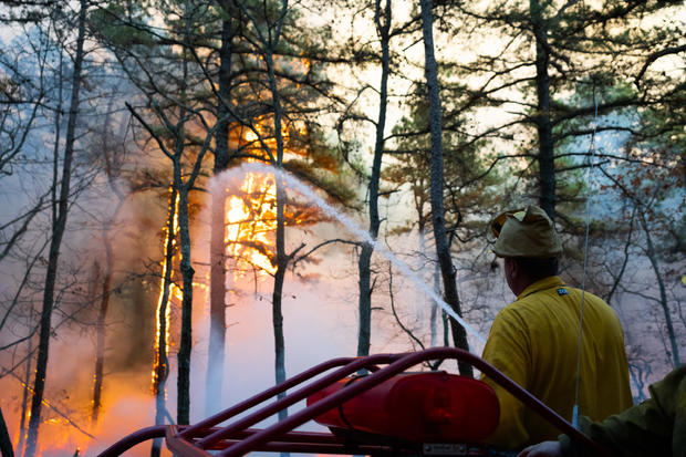 A firefighter holds a hose spraying water as flames are seen 