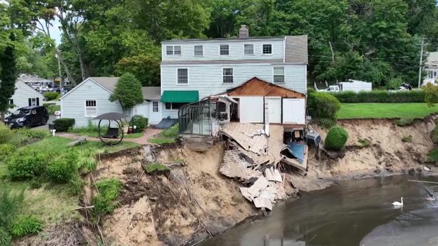 A home with a room that has partially collapsed and slid down a cliff into a pond. 