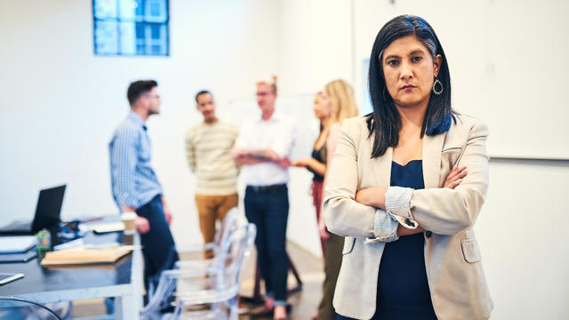 Male Coworkers Whispering Behind Back Of Unhappy Businesswoman In Office 