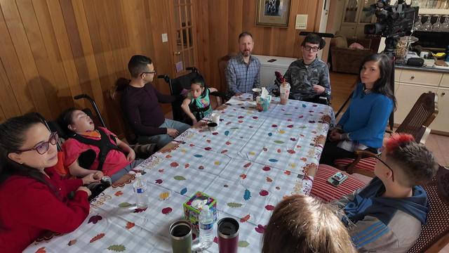 Five adults and four children, three of whom are in wheelchairs, sit around a kitchen table. 