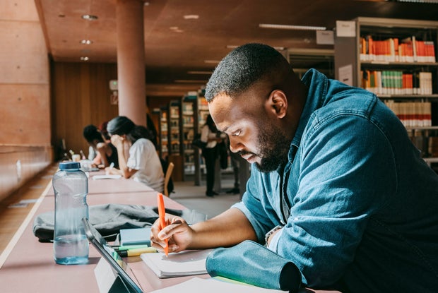 Student writing in notebook while sitting with tablet PC in college library 