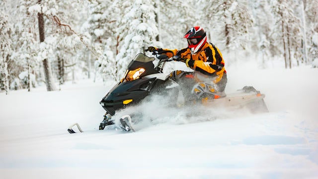 Human in the outfit of a racer in a multi-color jumpsuit and a helmet, driving a snowmobile by the deep snow surface on background of snowy forest. 