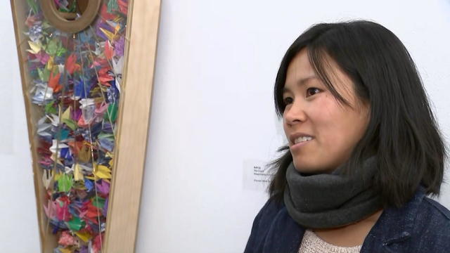 Anh Ly speaks to a reporter, art is hanging nearby -- a shadow box filled with origami 