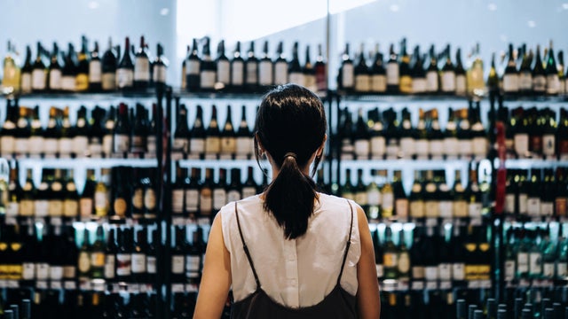 Rear view of young Asian woman grocery shopping for wines in a supermarket. She is standing in front of the liquor aisle and have no idea which wine to choose from