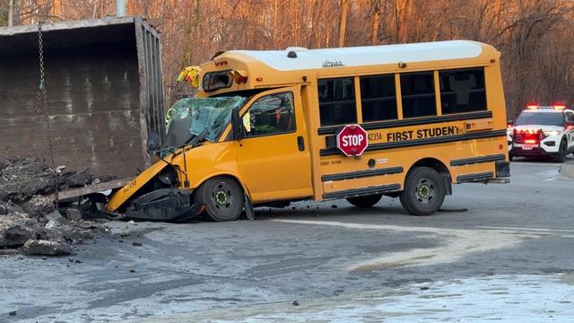 The corner of a dump truck crushes the front of a small school bus on a road. 