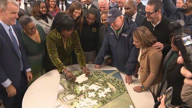 Mayor Cherelle Parker, surrounded by a crowd looking on, places a model building on a replica of the wellness center campus 