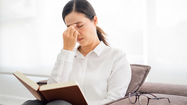 Lady suffering eyestrain reading book sitting on a couch in living room at home