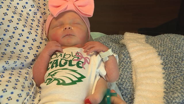 A newborn baby wearing a pink bow and a onesie that says "Baby Eagle" in her mother's arms 