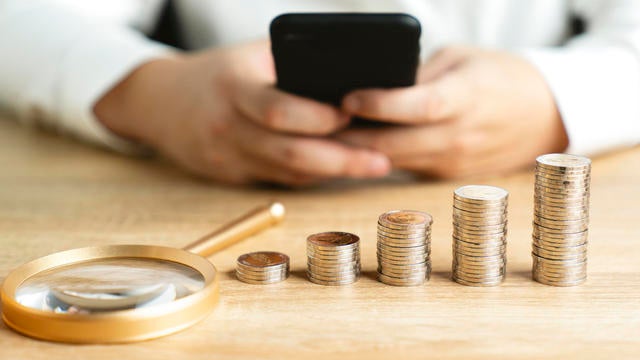 Businessman is opening data on his mobile phone to earn more money with magnifying glass and pile of money on the table. 