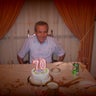 Chafic "Steve" Ezzeddine sits at a table with a birthday cake with candles in the shape of the number "70." 