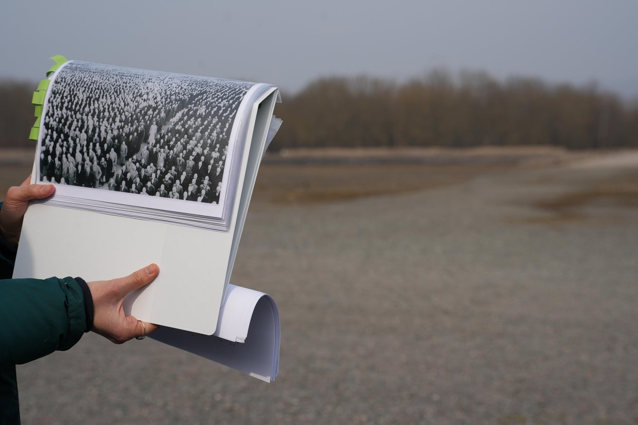 Rose Shafer, a volunteer at the Buchenwald Memorial, holds a historic photo of the roll call grounds packed with prisoners against the backdrop of the same space as it appears today