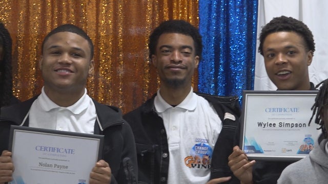 Three young men smile, two are holding up their certificates in this photo from the graduation ceremony 