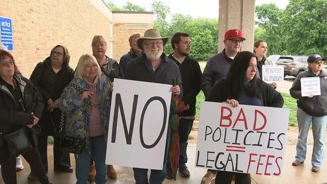 Parents and community members hold signs outside a school, one sign says NO and another says bad policies equals legal fees 
