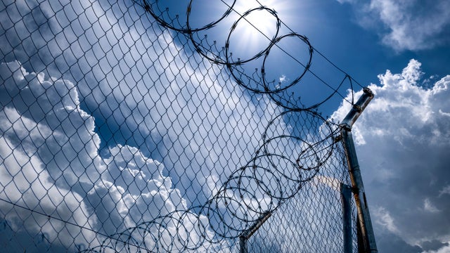 Close-up of metal fence with barbed wire over a dramatic sky with the sun on the horizon. 