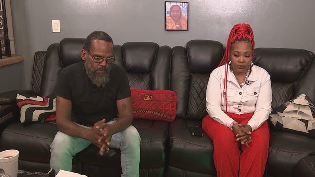 Isya Stanley's parents sit on the couch, a photo of Isya hangs behind them 