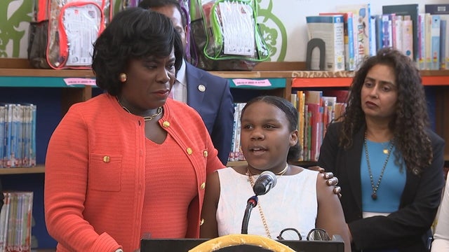 Mayor Cherelle Parker stands with a girl who is speaking during the bill signing, library shelves are in the background 