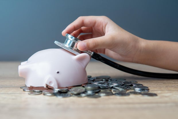 A hand holding a stethoscope listening to a broken piggy bank lying on a pile of coins.