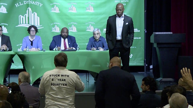 A man wearing a shirt reading "stuck with 8-year squatter" speaks to Mayor Eric Adams. 