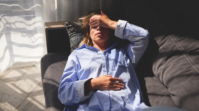 Woman exhausted and lying on couch. Hand on forehead, eyes closed. Indoors with natural light near a window with curtains. 