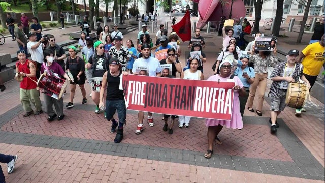 Demonstrators march behind a banner reading "Fire Jonathan Rivera." 