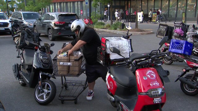 A grocery delivery worker loads bags of food into a container on the back of a moped. 