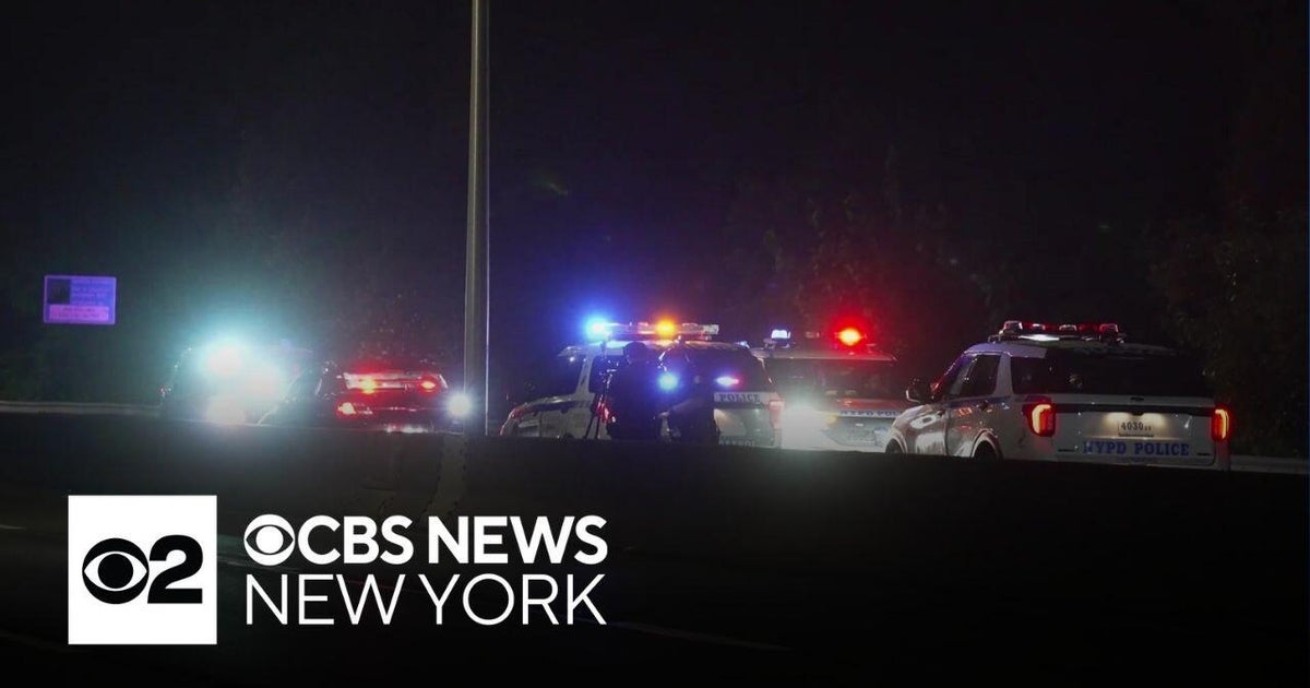 Multiple NYPD police SUVs and cars with red, blue and white flashing lights stopped on a dark roadway at night; a concrete median and a tall light pole are visible in the foreground.