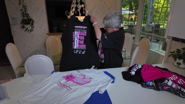 Dr. Ellen Mandel holds up a Race for the Cure t-shirt. A pile of other t-shirts sit on a table. 