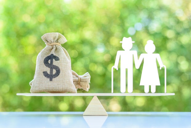  Balancing Retirement Income and Expenses. A miniature elder couple standing on a basic balance scale with US dollar money bag. Elderly face challenges in retirement. 