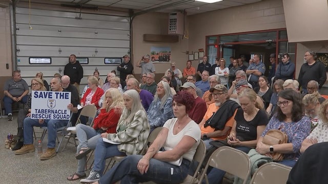 People sit in folding chairs in the firehouse during the meeting, one person has a sign that says Save the Tabernacle Rescue Squad 