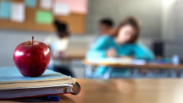 Apple and a pile of books and pens on teacher table in classroom. Focus on foreground. 