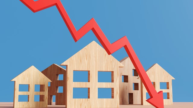 Red downward arrow in front of wooden house models on a wood table on a blue background. Illustration of the concept of falling prices of real estate properties 