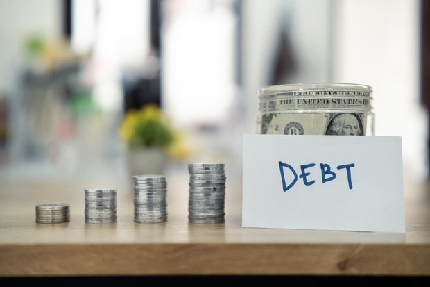 Close-up of stacked coins beside a note labeled debt with a jar filled with cash in the background, symbolizing financial burdens, savings, or economic planning issues.