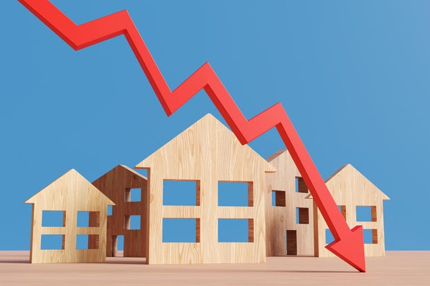 Red downward arrow in front of wooden house models on a wood table on a blue background. Illustration of the concept of falling prices of real estate properties 