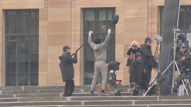 An actor dressed as Rocky, surrounded by a film crew, jumps at the top of the Philadelphia Art Museum steps 