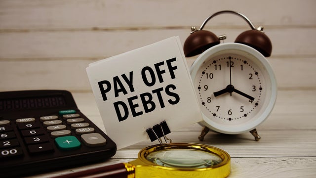 Pay Off Debts text on paper card with alarm clock, calculator and magnifying glass on wooden background 