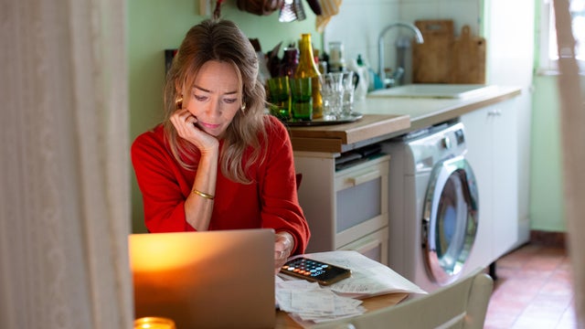 Woman sitting at kitchen table, using laptop and sorting through paperwork 