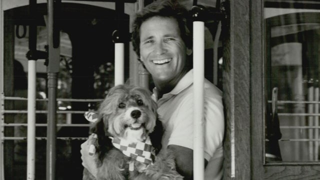 A man smiles while holding a dog that is wearing a bandana scarf 