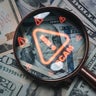 Magnifying glass focusing on scam warning symbol over dollar bills concept of financial fraud money laundering cybercrime and online scam alert for security awareness and finance protection 