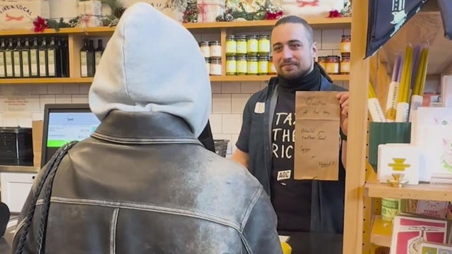 A man stands behind a cash register holding a sign that asks a single question 