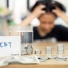 A woman sits at a table, looking distressed, with piled coins and a debt note. This scene expresses the concept of financial burden, economic challenges, and personal reflections on money management. 