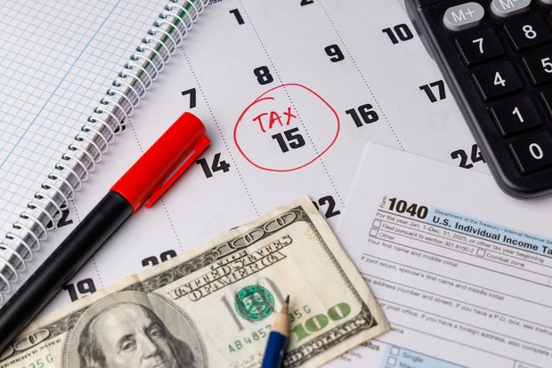 Close up of a calendar with a red circle on the 15th and color Form 1040 next to a hundred dollar bill and modern office supplies shot. 