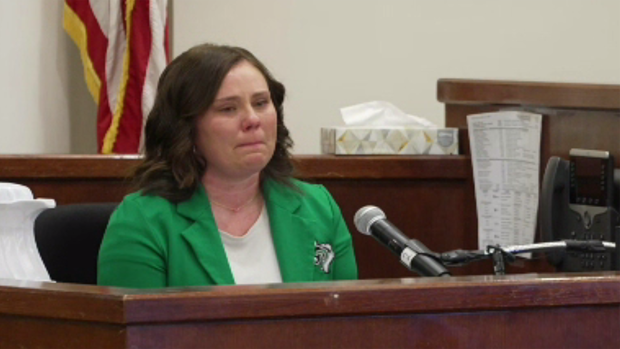 Lindsey Thompson seated at brown court witness stand, wearing a green blazer and white blouse. She has brown hair and gray eyes. 