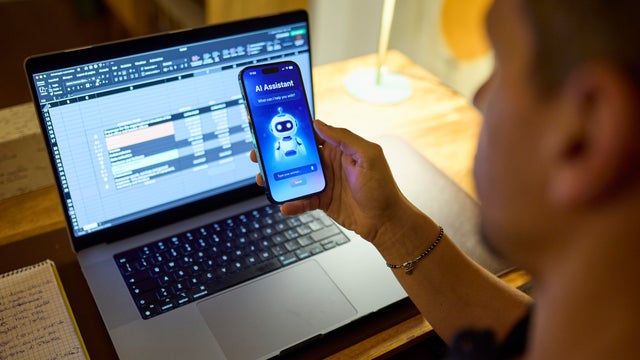 Man using AI assistant on smartphone while working on laptop at home desk at night 
