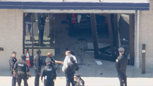 Helicopter photo of the broken door at a police station, a blue car is inside the building, police and others are standing outside 