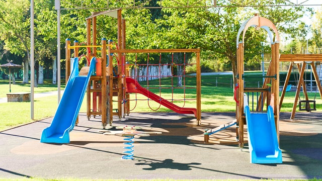 A playground with slides in blue and another in red, wooden structure and cement floor, roofed, with trees and grass around it, during the day. 