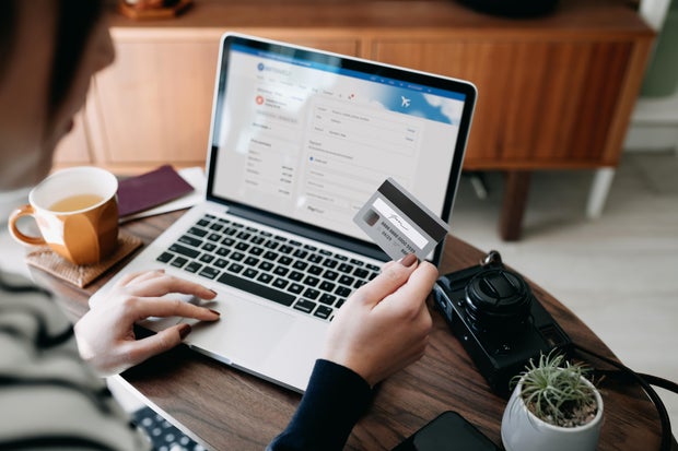 Over the shoulder view of young Asian woman shopping online for flight tickets on airline website with laptop, entering credit card details to make mobile payment at home. Camera and passport on the table. Travel planning. Booking a holiday online 