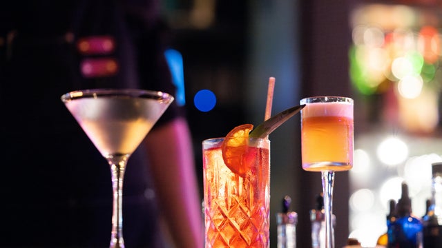 Three vibrant cocktails sit in a dark, trendy bar 