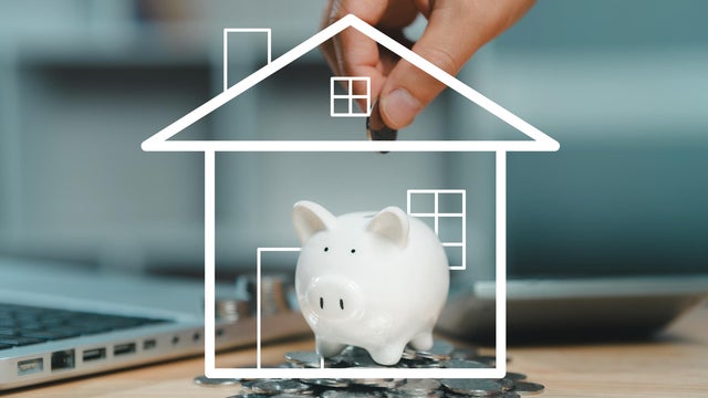 A hand dropping a coin into a piggy bank encased in a white line-art outline of a house, symbolizing the concept of saving money for a home purchase or mortgage. 