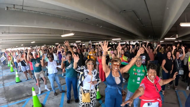 Potential contestants for The Price is Right wave to the camera from their line in a parking garage 