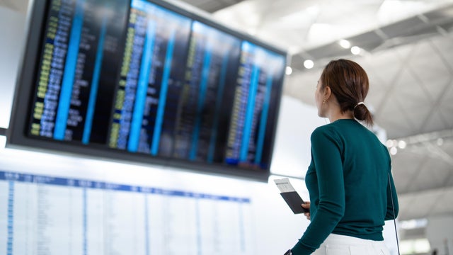 Young Asian woman with passport and boarding pass, looking up and checking her flight schedule from the Arrival Departure Board at airport terminal. Young female tourist ready for a trip. Business travel. Travel and vacation concept 